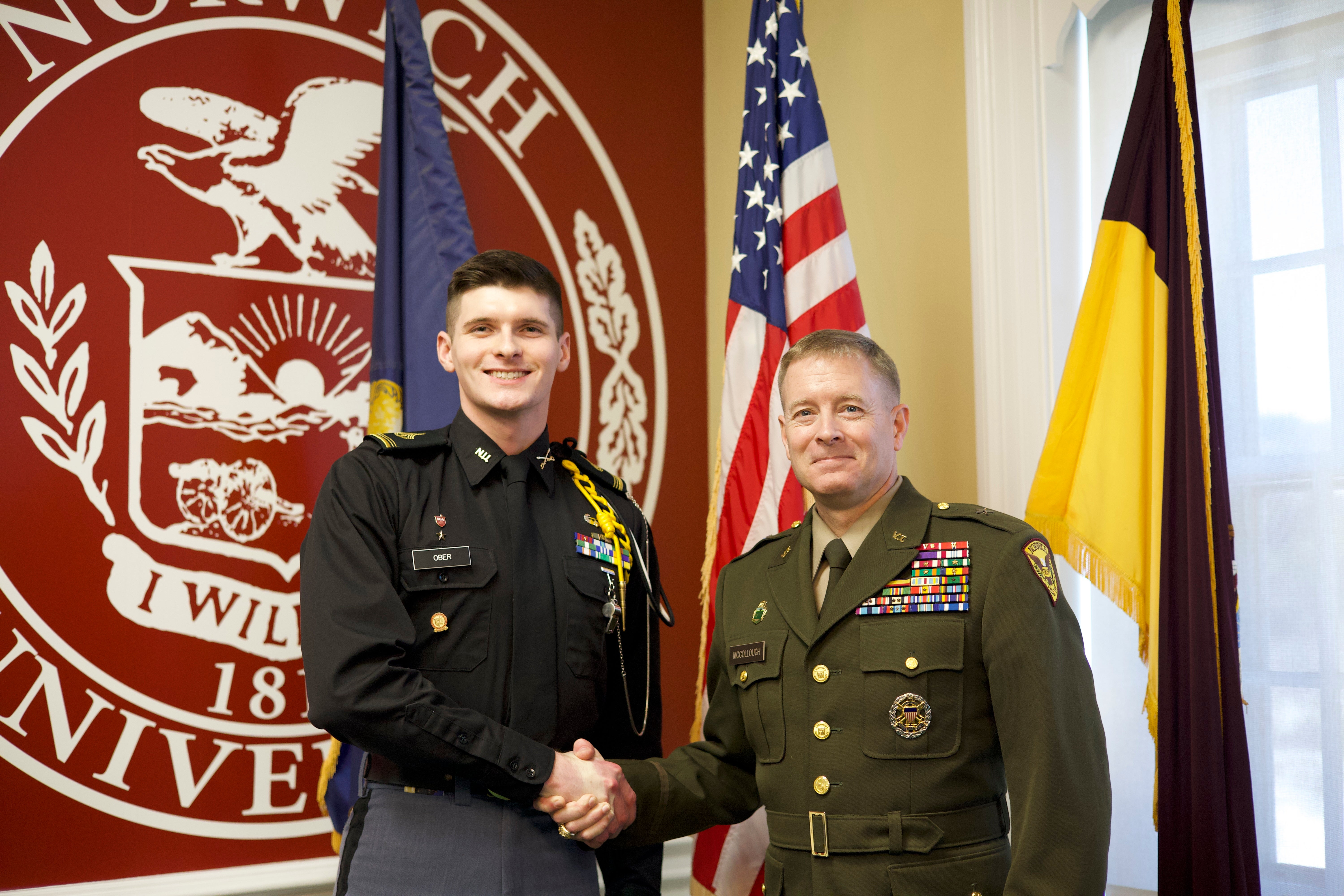 BG William F. McCollough, VSM, Commandant and Vice President for Student Affairs, Norwich Class of 1991 and C/1SG Matthew S. Ober '26 shaking hands in front of the Norwich University logo