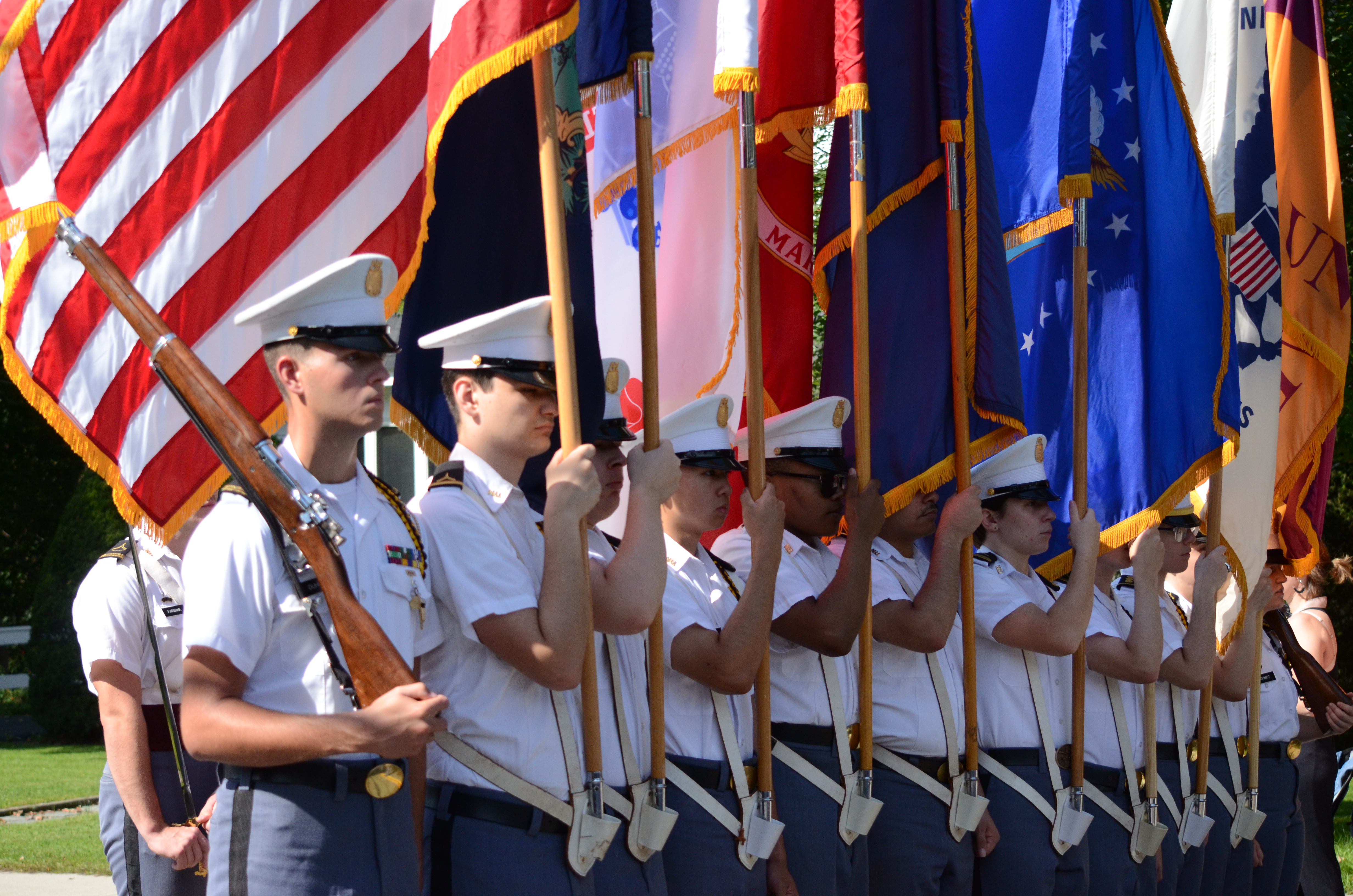 Cadets in white uniforms stand in a row, holding colorful flags and rifles during an outdoor ceremony.