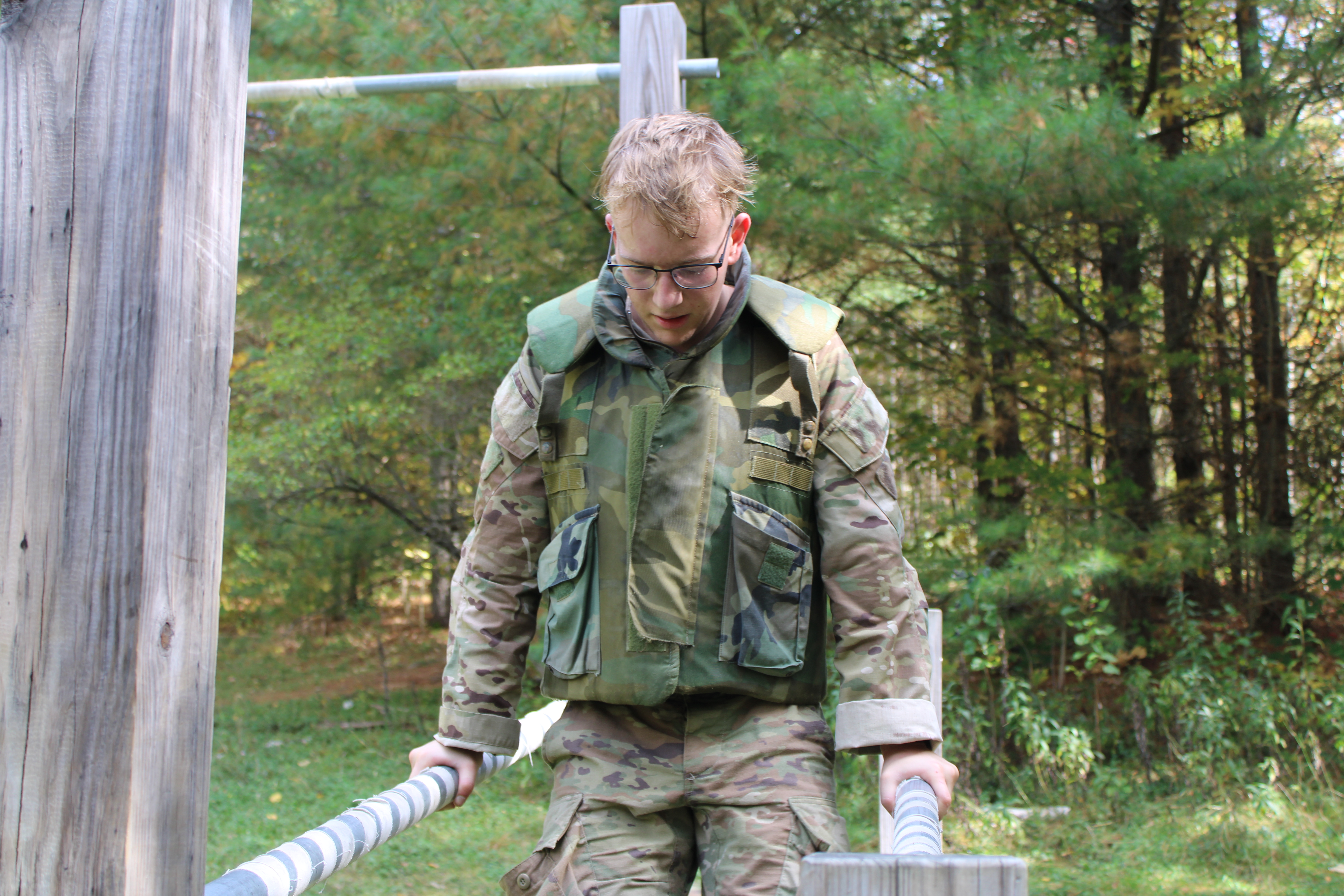 A person in military camouflage uniform performing an exercise on parallel bars in an outdoor obstacle course. The background features a wooded area with green foliage.