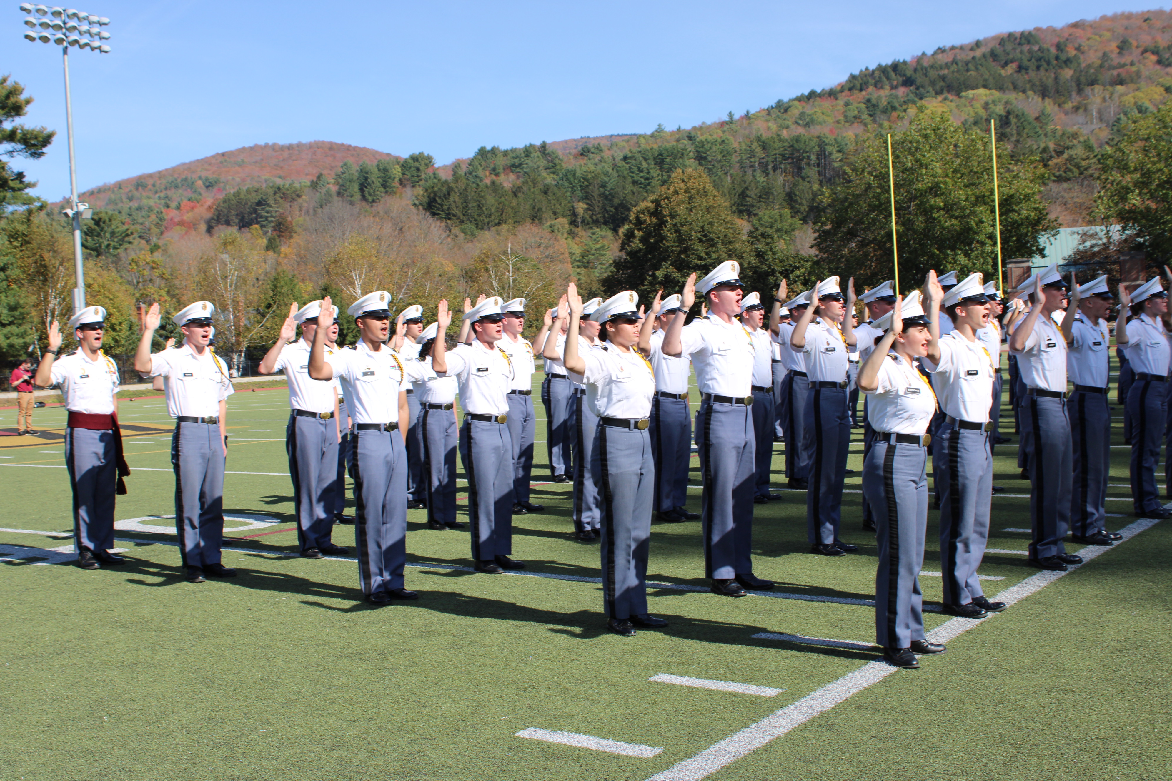Cadets in uniform stand in formation on a field with raised hands during a ceremony.