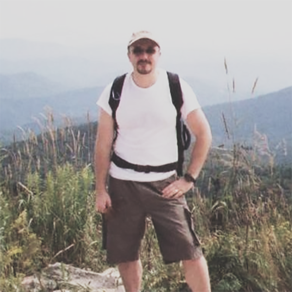 A man stands in hiking gear atop a mountain with a scenic view in the background.