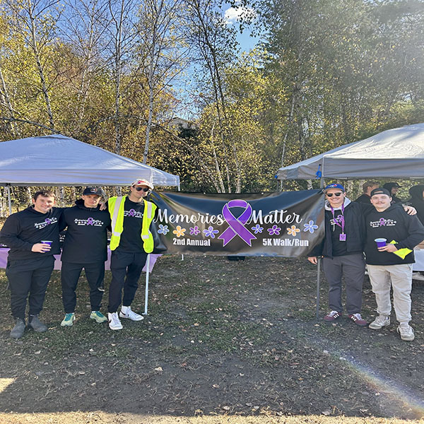 A group of five people are standing together outdoors in front of a banner that reads “Memories Matter – 2nd Annual 5K Walk/Run.” The banner features a large purple awareness ribbon surrounded by flowers. The participants are dressed warmly in black “Memories Matter” shirts and casual athletic wear, with one person wearing a bright yellow safety vest. They’re standing between two canopy tents on a clear, sunny day with trees showing fall foliage in the background.
