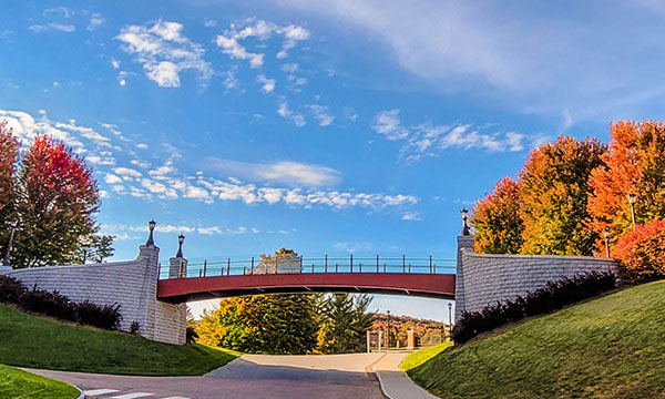A brick and stone bridge crosses over a road, surrounded by green grass and colorful autumn trees under a blue sky.