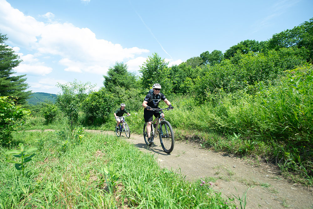 Two people ride mountain bikes on a dirt path through green, leafy trees under a blue sky with scattered clouds.