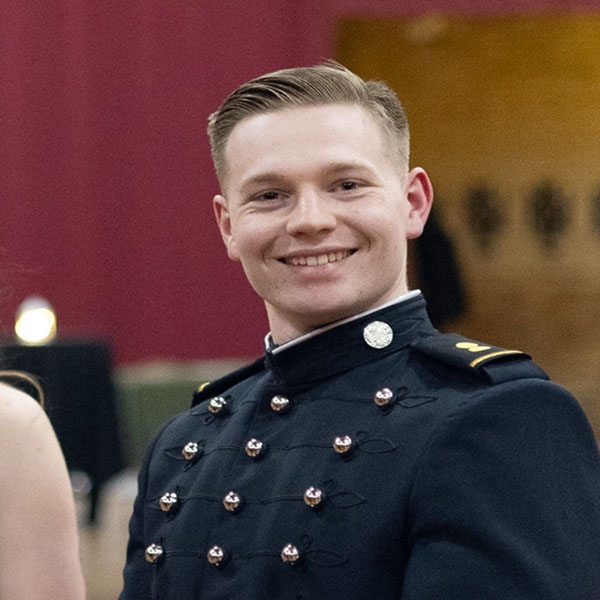 A smiling cadet in a formal dark military uniform with shiny buttons poses indoors, standing against a softly lit background.