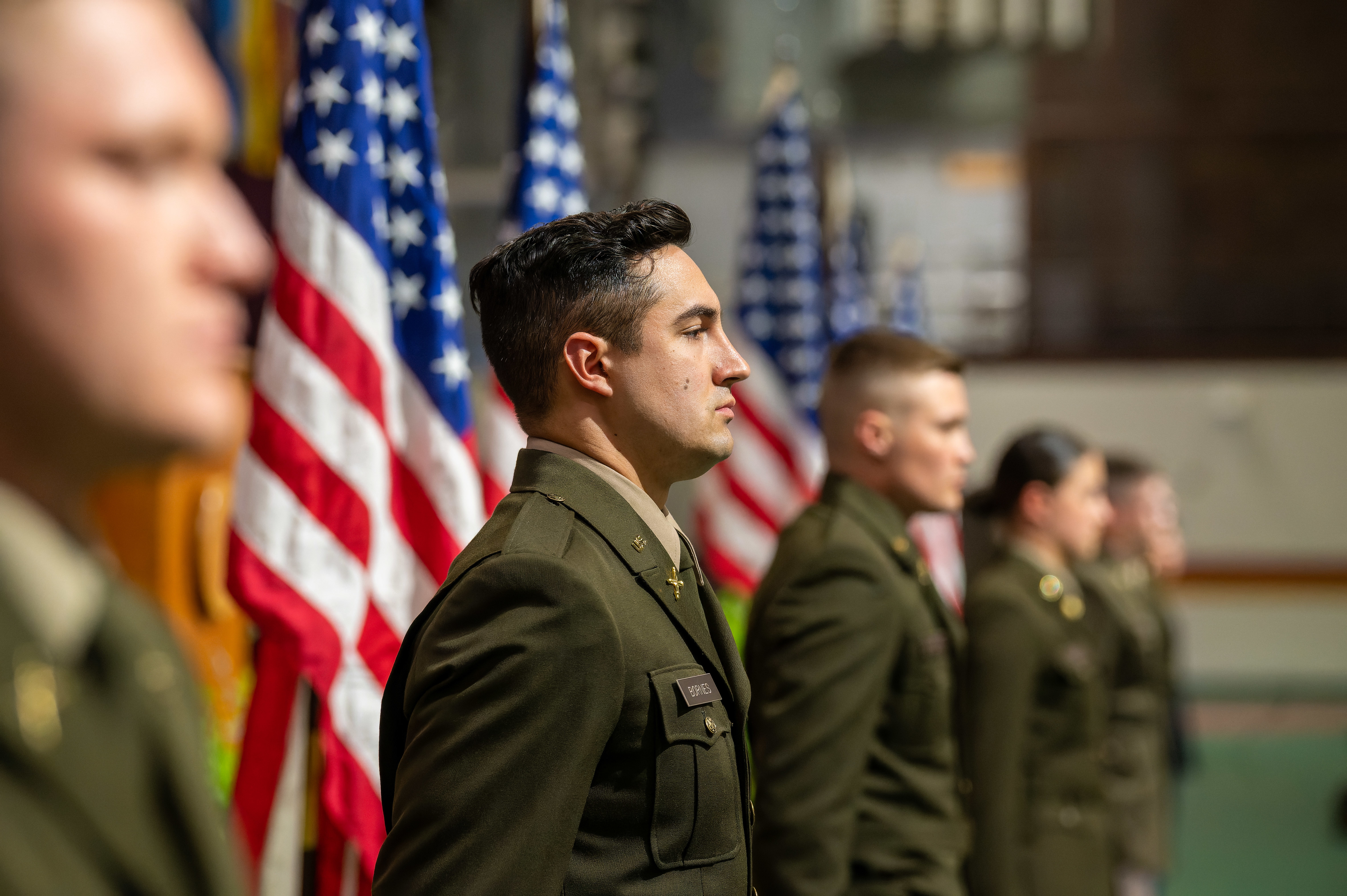 Uniformed cadets stand in a line with American flags in the background.