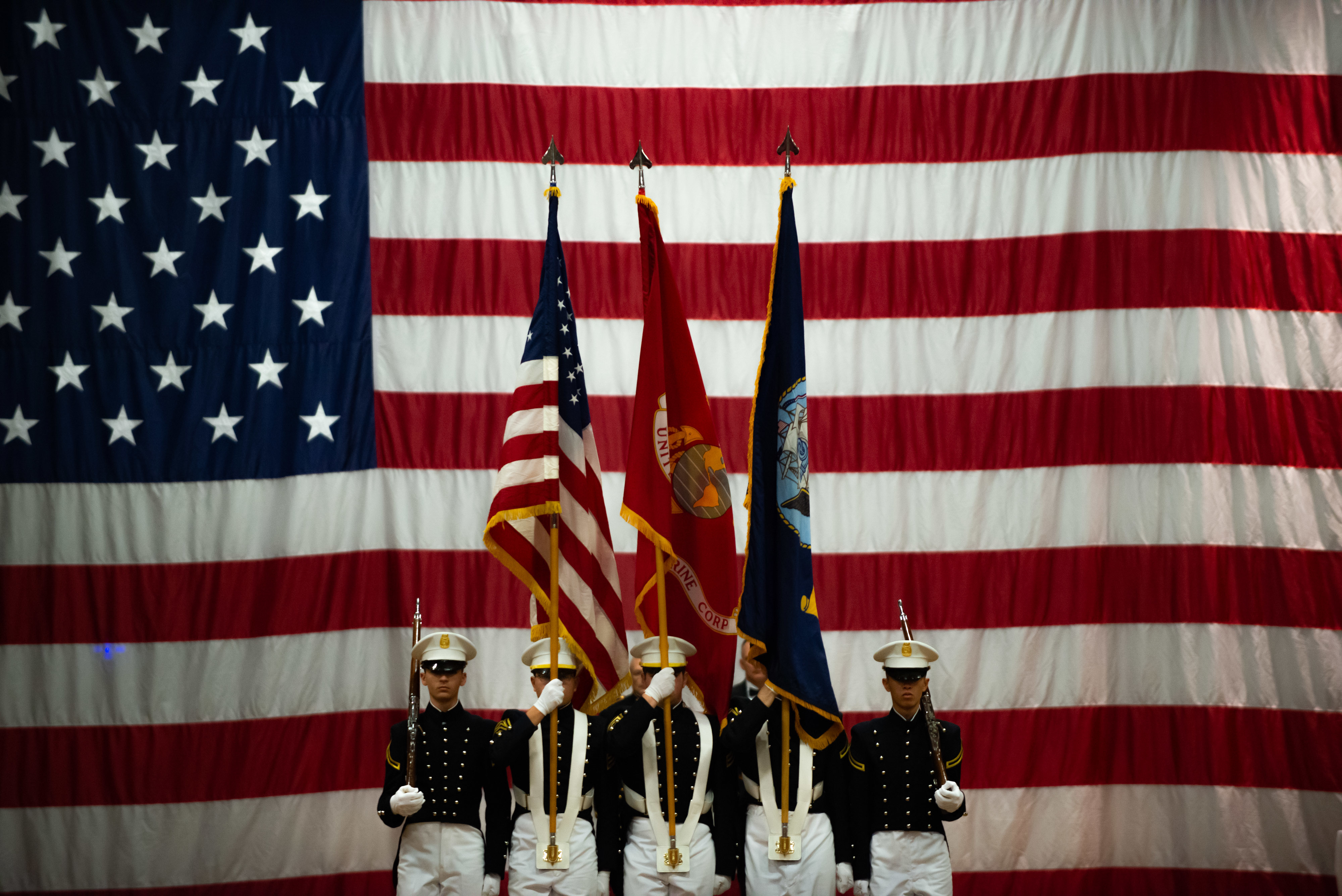 Military honor guard stands in front of a large American flag, holding three flags.