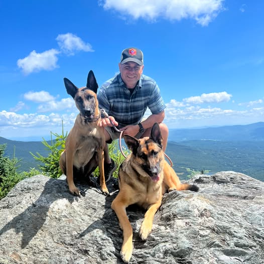 A man standing on a mountainside smiles, posing behind his two dogs.