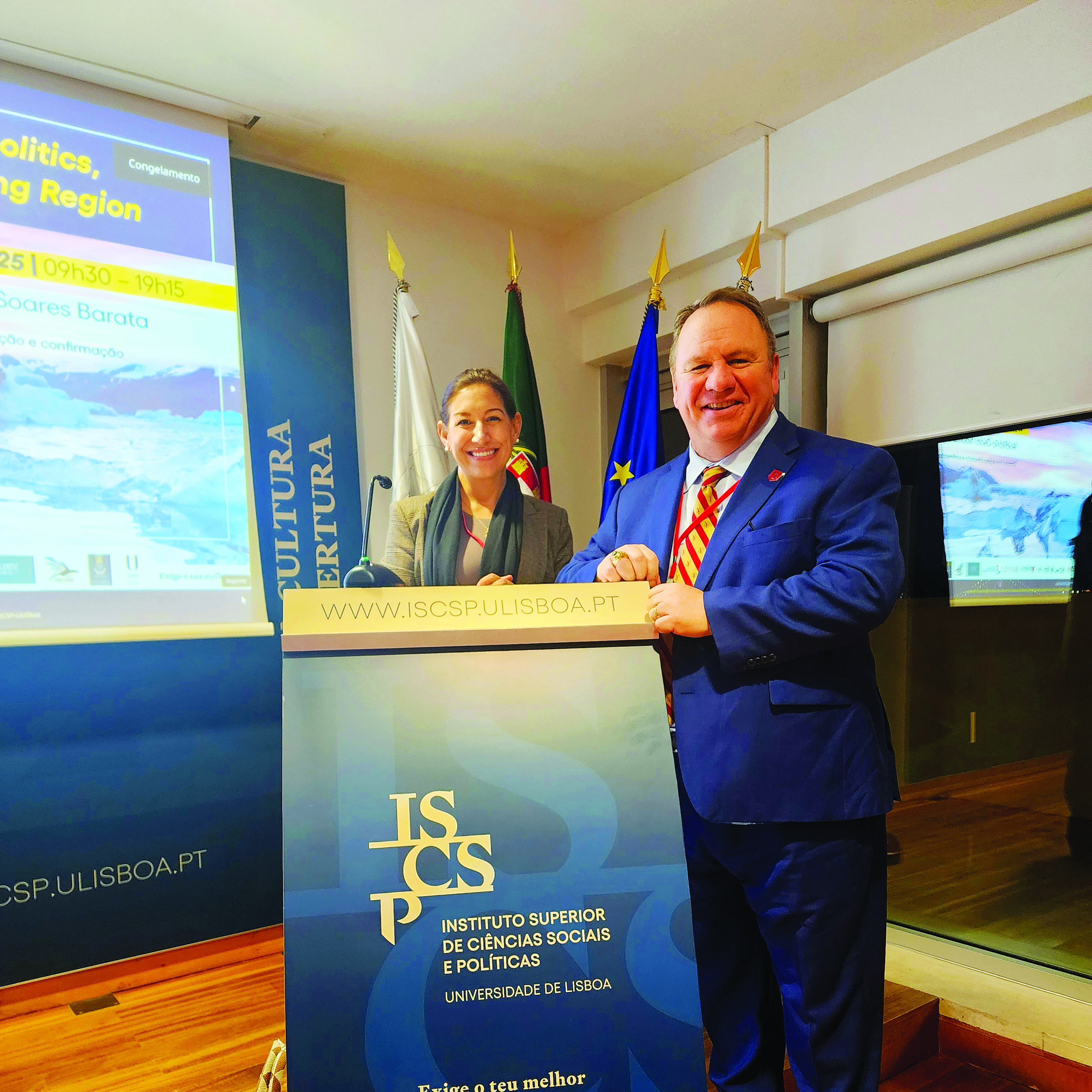 Two people stand at an ISCTE podium in a conference room with flags and presentation screens behind them.