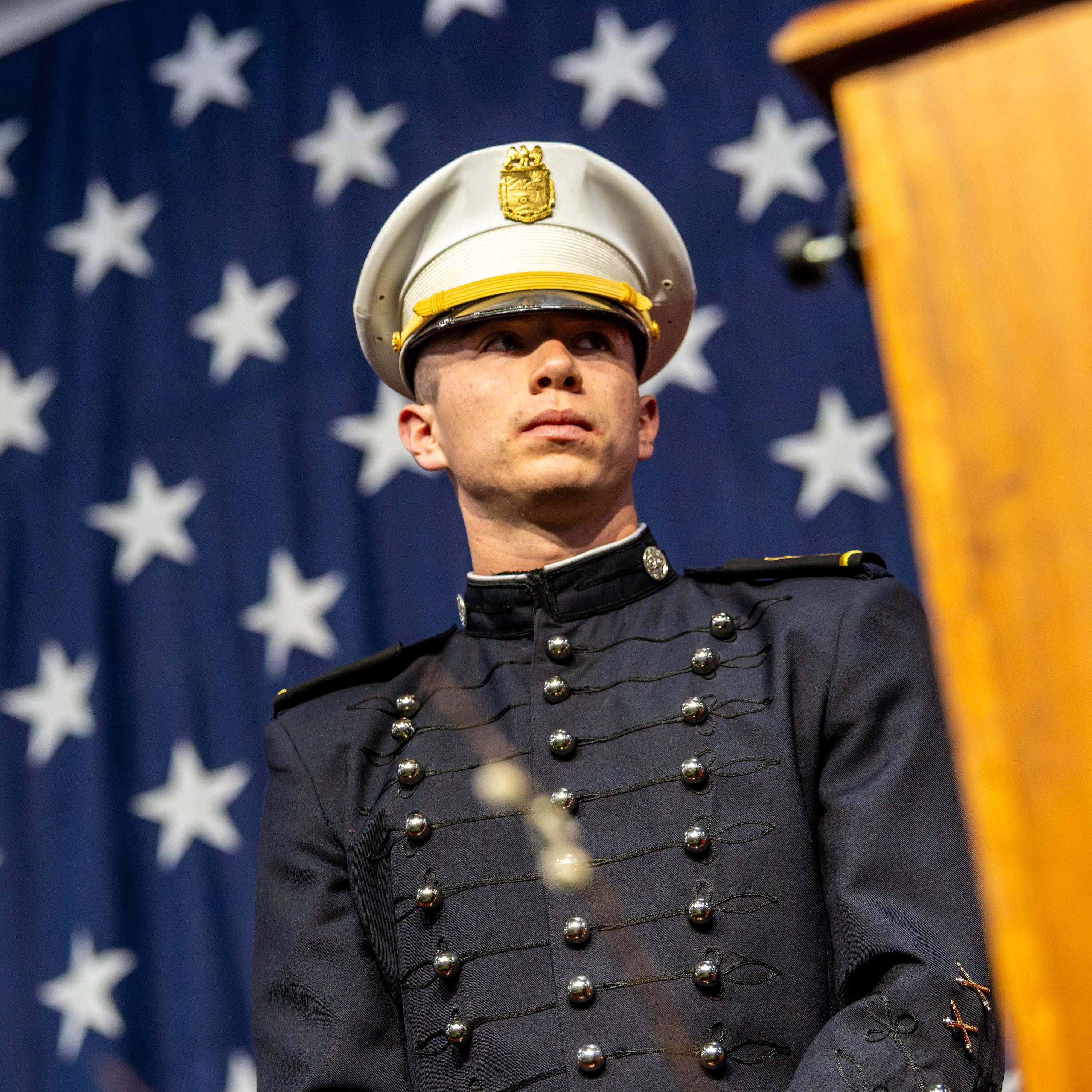 Cadet in dress uniform stands before large American flag during ceremony.