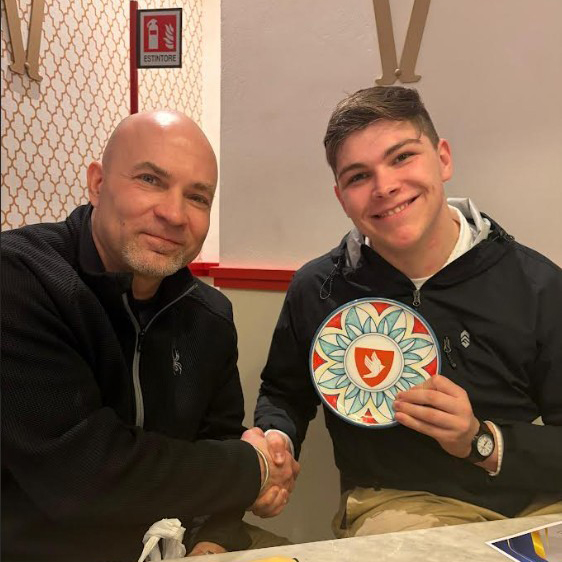 Two people sit at a table shaking hands, while one holds a circular artistic tile.