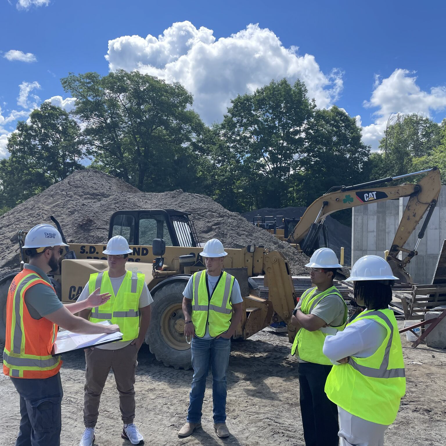 Students in hard hats and safety vests receive instruction at a construction site with heavy equipment.