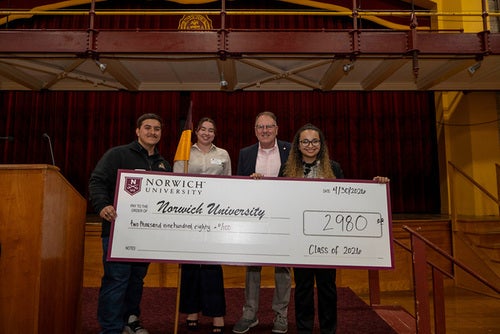 Four people hold a ceremonial check for $2,980 from Norwich University labeled Class of 2026 on a stage.
