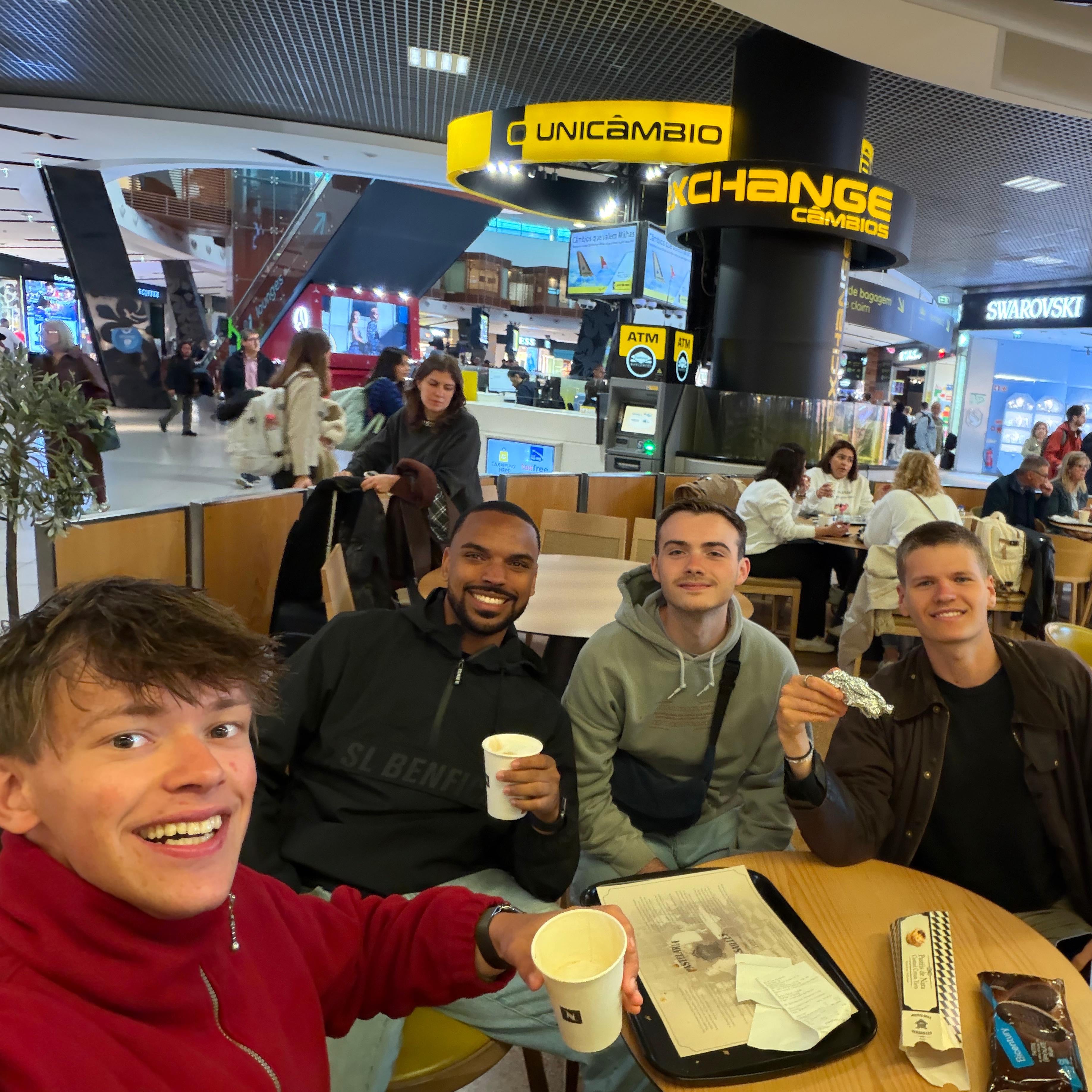 Four people sit around a food court table holding drinks and smiling for a selfie in a shopping mall.