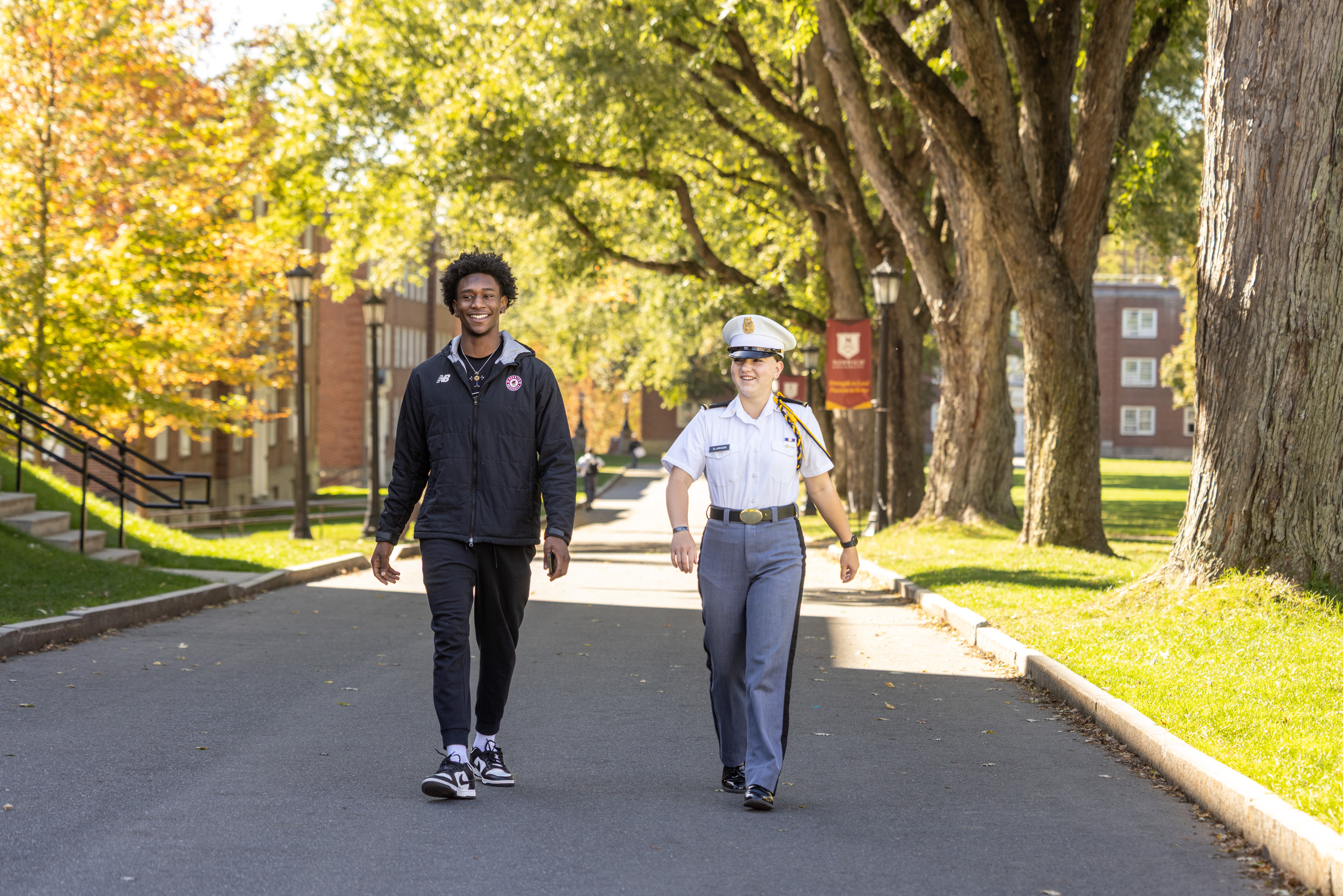 Student and cadet walk together along a tree-lined campus road.