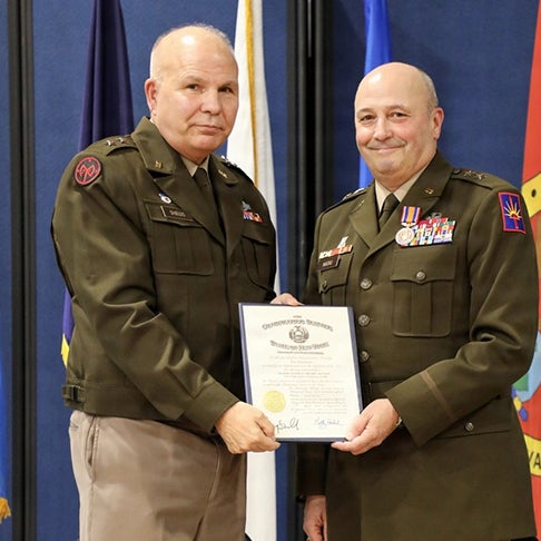 Two service members in dress uniforms hold a framed certificate in front of flags.