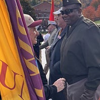 A person holding a yellow and purple flag speaks with a person in a military-style uniform at an outdoor gathering.