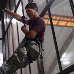 Cadet wearing a safety harness climbs an indoor obstacle while holding a rope.
