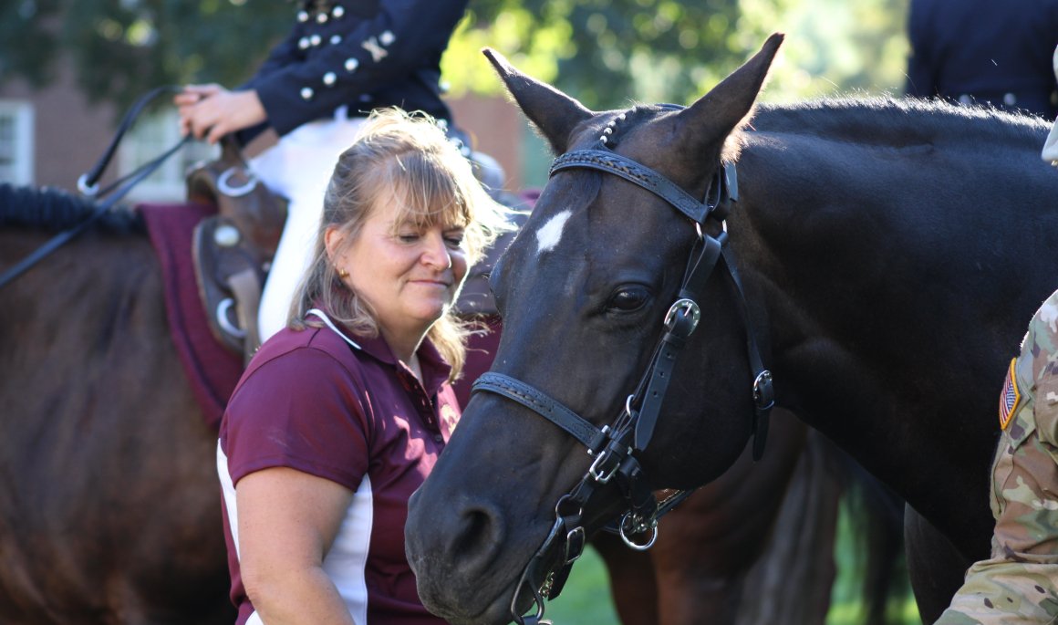 Woman standing beside a dark horse as riders sit mounted in the background.