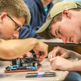 Two Norwich University students work with focused concentration, assembling electronic components on a circuit board at a classroom table.