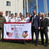 A group photo of the Olmsted trip group, holding a branded Norwich/Olmsted flag in uniform in front of military academy.