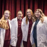 Four individuals stand together smiling, dressed in maroon scrubs and white coats embroidered with “Norwich University School of Nursing.” The backdrop of rich red curtains and stage lighting gives the setting a formal and celebratory tone.