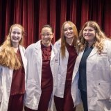 Four individuals stand together smiling, dressed in maroon scrubs and white coats embroidered with “Norwich University School of Nursing.” The backdrop of rich red curtains and stage lighting gives the setting a formal and celebratory tone.