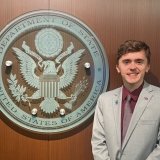 A student in a grey suit stands next to the Department of State logo.