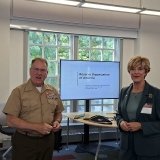 Two individuals formally dressed stand in front of a presentation screen.