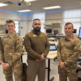 Two cadets and an instructor stand in a computer lab classroom.