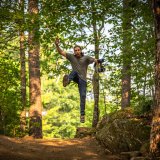A person jumps mid‑air on a forest trail holding a camera with sunlight filtering through the trees.