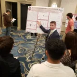 Student presents a research poster to a group at an indoor academic conference.