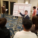 Student presents a research poster to a group at an indoor academic conference.