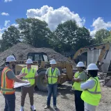 Students in hard hats and safety vests receive instruction at a construction site with heavy equipment.