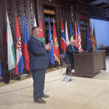A speaker addresses an audience beside seated panelists with national flags displayed across the stage.