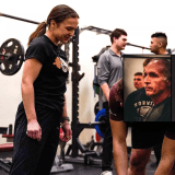 Strength coach watches a weightlifting session in a gym with several people and exercise equipment.