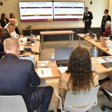 Presenter speaks to attendees seated around tables as two screens display a legacy fleet presentation.