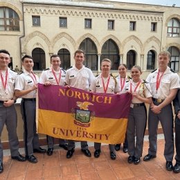 Norwich cadets stand shoulder-to-shoulder holding a Norwich flag, flanked on each end by Norwich faculty. They are standing in a courtyard with a building in the background.