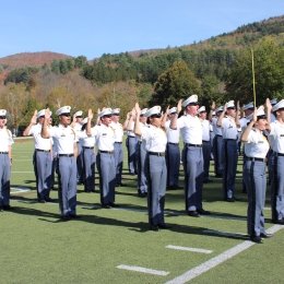 Cadets in uniform stand in formation on a field with raised hands during a ceremony.