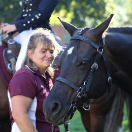 Woman standing beside a dark horse as riders sit mounted in the background.