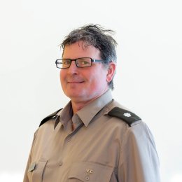 Person wearing glasses and a gray uniform shirt with shoulder insignia poses against a plain light background.