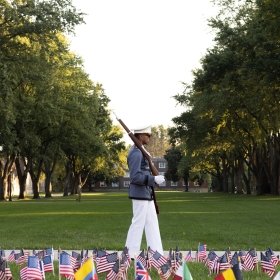 Flags are planted in the ground on the Upper Parade Ground immediately in the foreground; just behind them, a cadet walks along the Tour Strip with a rifle over his right shoulder, and a view of the Upper Parade Ground behind him.