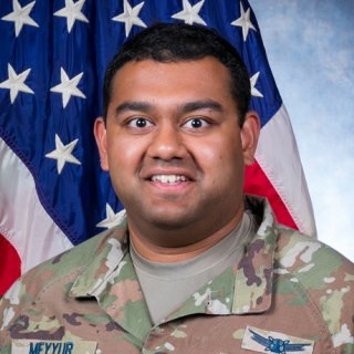 U.S. Space Force officer in camouflage uniform with name tape "Meyyur" stands before an American flag, displaying rank insignia and mission patches.