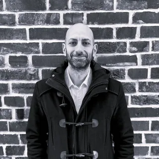 Man in a dark coat standing in front of a brick wall.