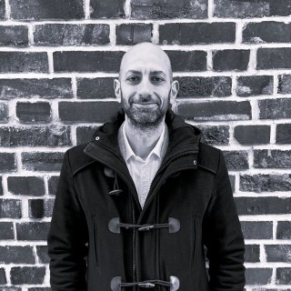 Man in a dark coat standing in front of a brick wall.