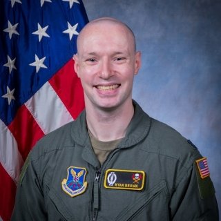 Smiling man in a green flight suit stands in front of a U.S. flag, with Air Force insignia and a name patch reading “Ryan Brown” on his uniform.