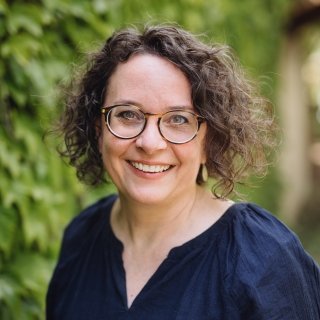 Woman with curly hair and glasses smiling, standing outdoors with greenery in the background.