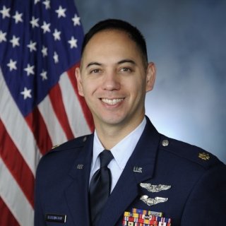 U.S. Air Force officer in dress blue uniform stands in front of the American flag, displaying aviation wings and a row of colorful service ribbons.