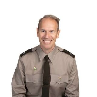 Individual wearing a tan military uniform shirt with dark brown shoulder epaulets, a dark brown tie, and two pins on the chest pockets against a white background.