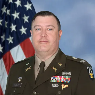 U.S. Army officer in dress uniform with name tag reading Dudley, various ribbons, badges, and insignia, standing in front of an American flag and blue backdrop.